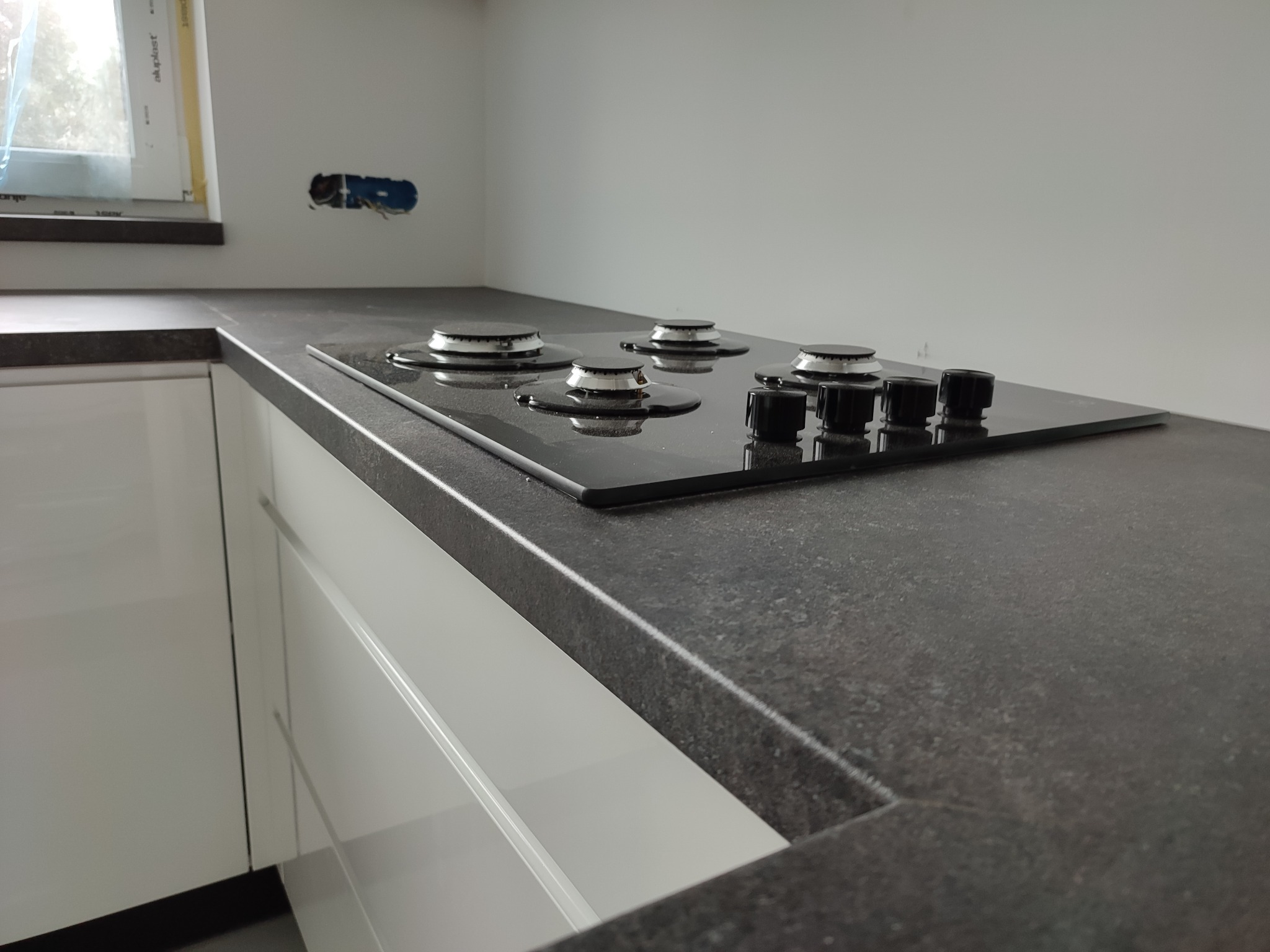 Szary, nakrapiany blat kuchenny z zamontowaną czarną płytą gazową z czterema palnikami, białe szafki kuchenne w tle, widoczne gniazdko elektryczne w ścianie.
