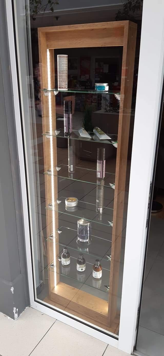 Wysoka, przeszklona witryna z drewnianą ramą i oświetleniem LED, prezentująca kosmetyki na szklanych półkach.