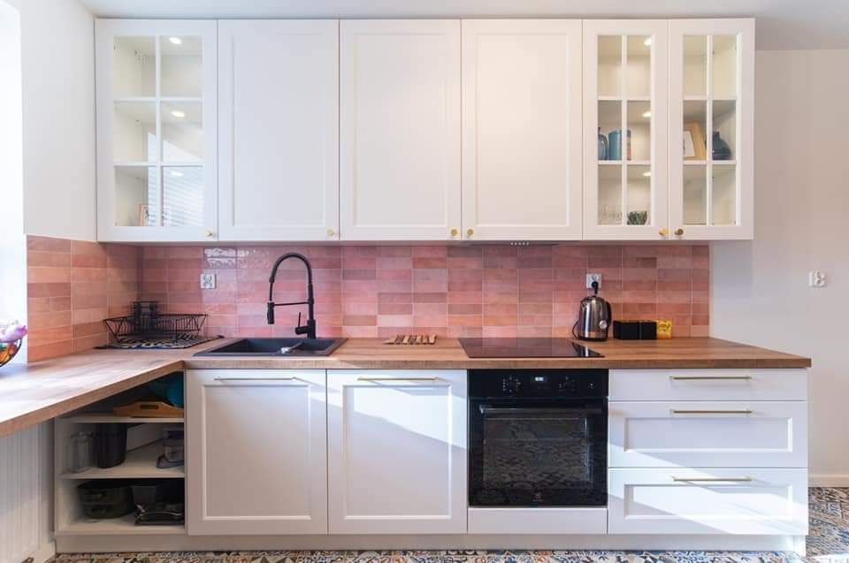 Biała zabudowa kuchenna z drewnianym blatem i różową mozaiką na ścianie, czarny zlew i armatura, szklane witryny górnych szafek z podświetleniem, wzorzysta podłoga.