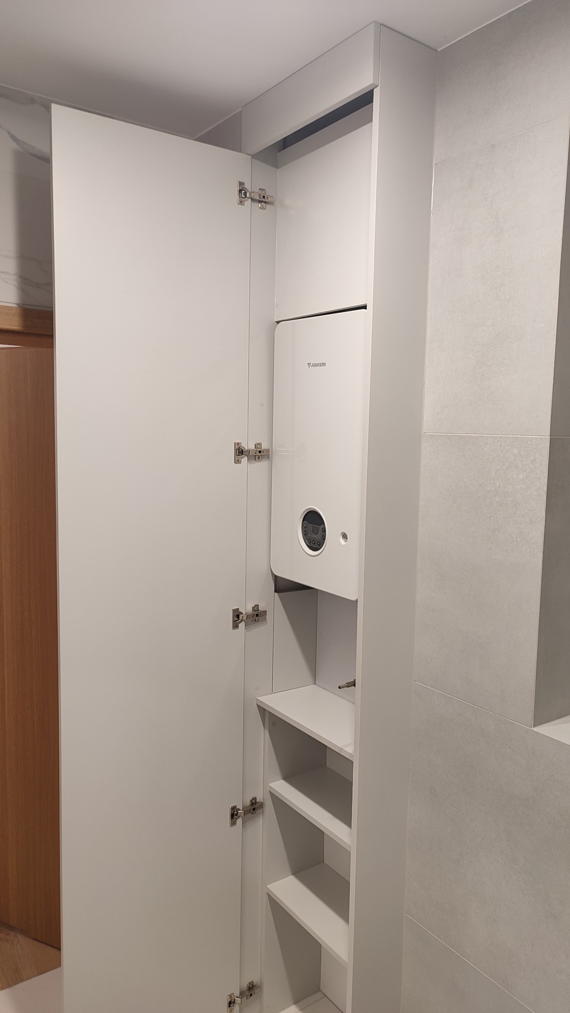 Biała szafa wnękowa z otwartymi drzwiami, wewnątrz zamontowany piec grzewczy Junkers i półki, widoczne zawiasy.