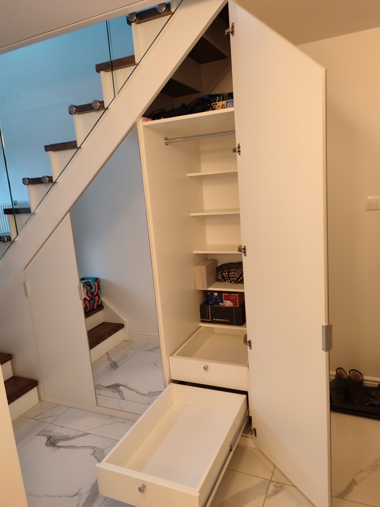 Biała szafa z otwartymi drzwiami i wysuniętą szufladą, zintegrowana ze schodami z szklaną balustradą, na marmurowej podłodze.