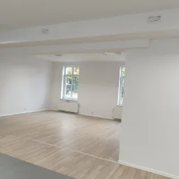 Pusty, odnowiony pokój z panelami podłogowymi o wzorze drewna, białymi ścianami, grzejnikami pod oknami i widocznymi kratkami wentylacyjnymi na suficie.