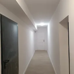 Długi, wąski korytarz po remoncie z jasnymi ścianami, panelami podłogowymi w odcieniach beżu i białymi drzwiami na końcu; po lewej stronie widoczna metalowa, ciemna i brudna obok wyremontowanego...