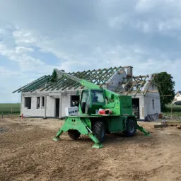 Wznoszenie domu murowanego: surowy stan otwarty z widocznym szkieletem dachu, wspierany przez zielony podnośnik teleskopowy Merlo na piaszczystym placu budowy.