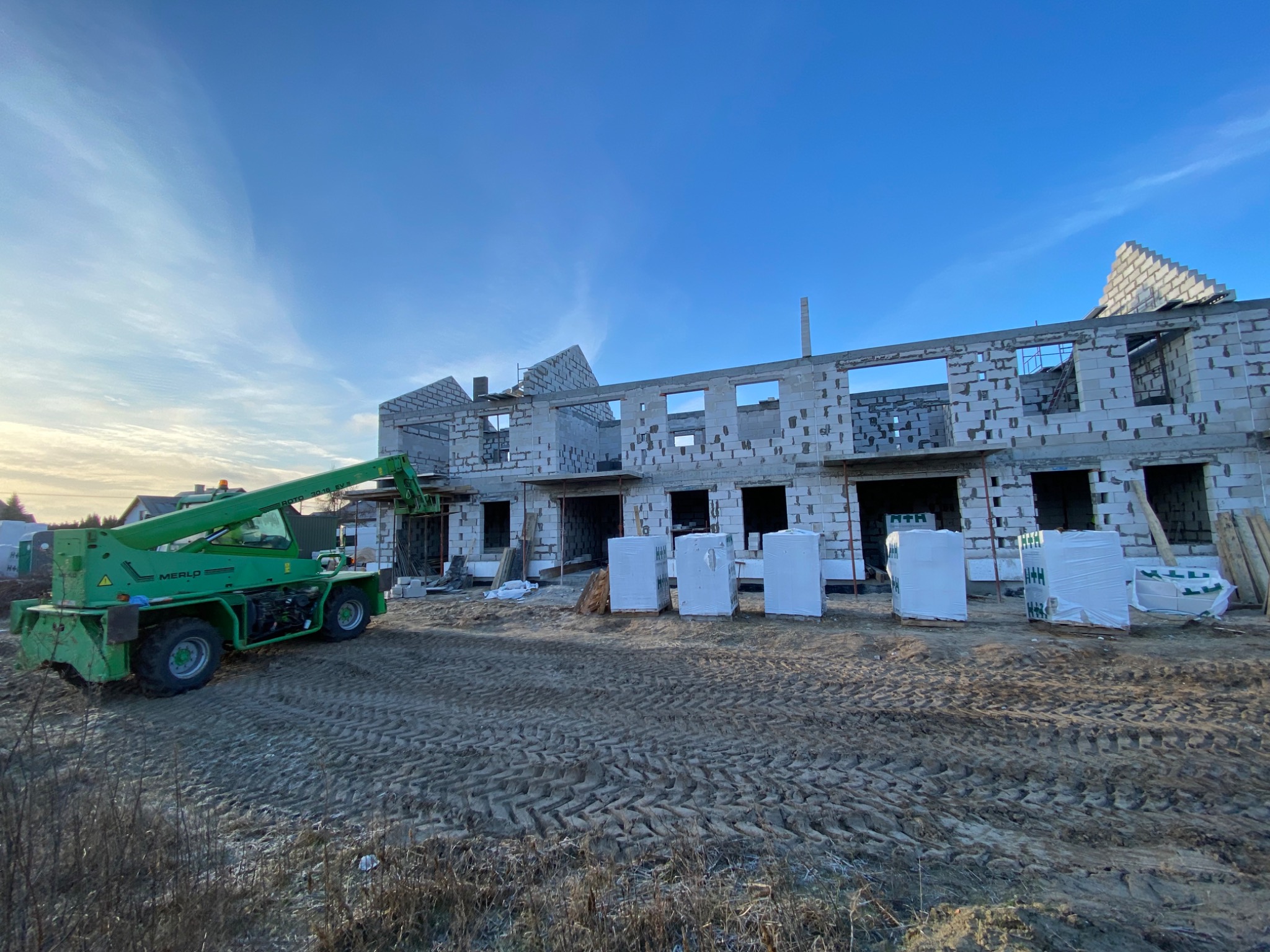 Wznoszony budynek z bloczków, widoczny szkielet konstrukcji, palety z materiałami budowlanymi i zielony podnośnik teleskopowy Merlo na placu budowy w słoneczny dzień.