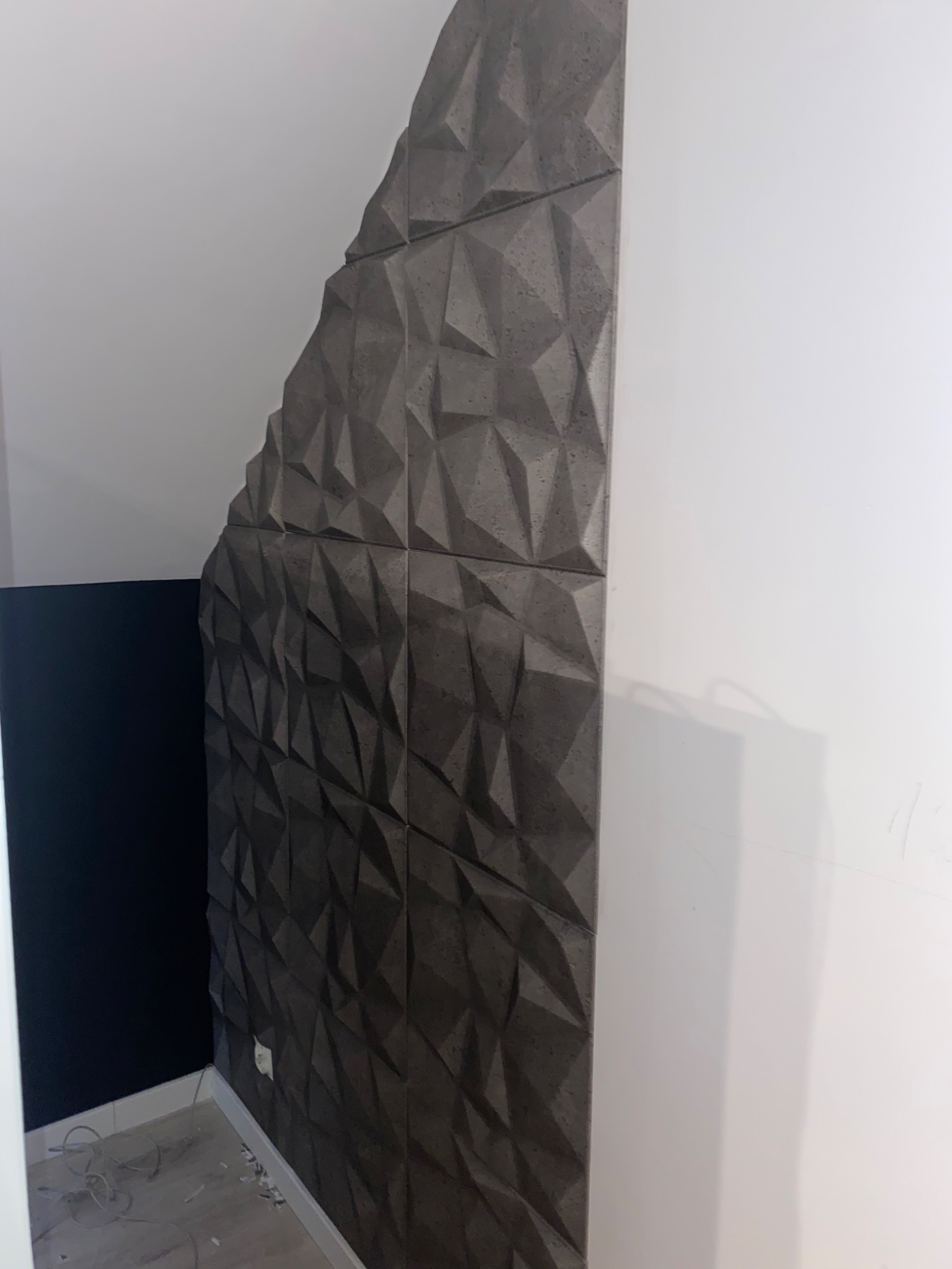 Fragment ściany z dekoracyjnych, trójwymiarowych paneli w odcieniach szarości, tworzących geometryczny wzór, przylegający do białej ściany i czarnego fragmentu ściany, widoczne przewody na podłodze.