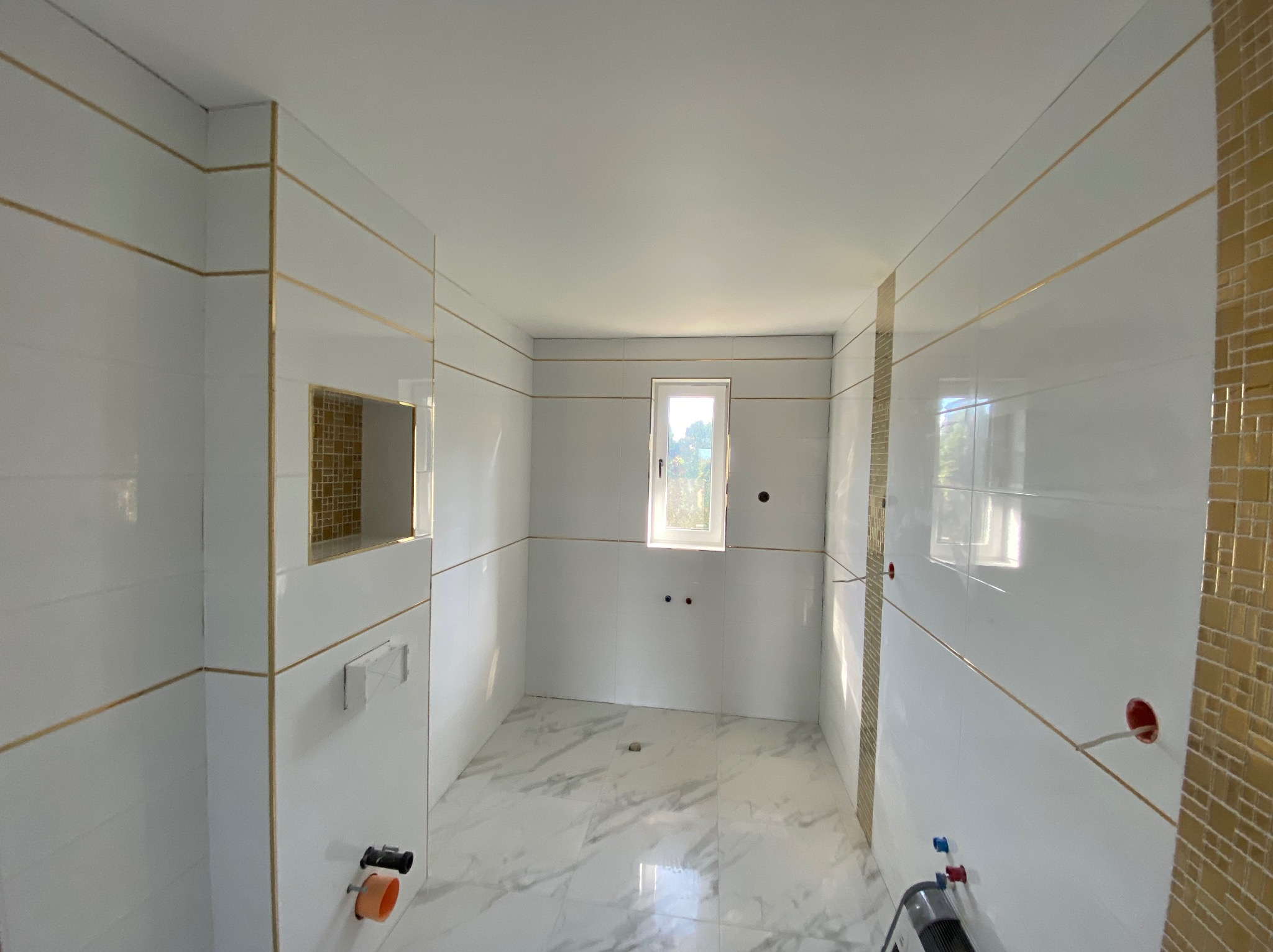 Wnętrze łazienki w trakcie remontu z białymi płytkami z delikatnymi złotymi akcentami, widoczne przygotowane instalacje wodne i fragment mozaiki w niszy.