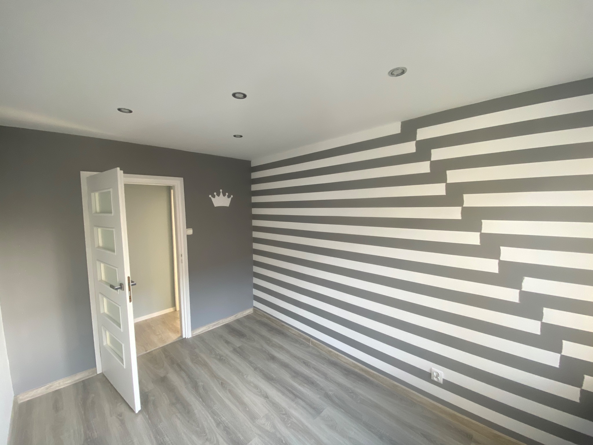 Pokój po malowaniu ścian w szaro-białe pasy z efektem schodkowym, szara ściana z naklejką korony, otwarte białe drzwi z przeszkleniami, szara podłoga imitująca drewno, oświetlenie sufitowe punktowe.