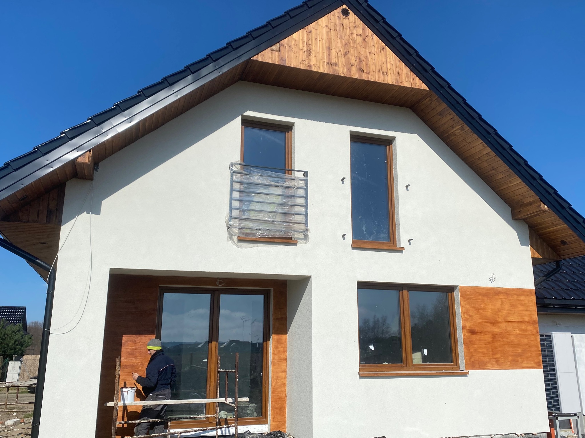 Pracownik na rusztowaniu maluje elewację domu jednorodzinnego w Grudziądzu, z widocznym drewnianym wykończeniem szczytu i okien.