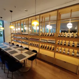 Sala restauracji z długim stołem nakrytym dla gości, oświetlonym wiszącymi lampami, z drewnianą zabudową regałową pełną butelek wina w tle.