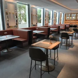 Wnętrze restauracji z zabudowanymi lożami w odcieniach brązu, stolikami z jasnymi blatami i krzesłami barowymi na tle dużych okien i regału z butelkami.