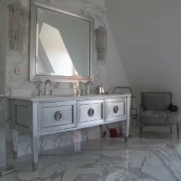 Elegancka łazienka w odcieniach bieli i srebra, z marmurową posadzką i ścianami, wyposażona w umywalkę z szafką z trzema szufladami, duże lustro, kryształowe kinkiety i dekoracyjne krzesło...