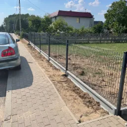Nowo zamontowane, ciemnoszare ogrodzenie panelowe z podmurówką, widoczne z perspektywy chodnika z kostki brukowej, z fragmentem srebrnego samochodu po lewej stronie i domem w tle.