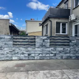 Nowoczesne ogrodzenie z betonowych bloczków o marmurkowym wzorze, uzupełnione poziomymi, szarymi przęsłami, na tle zabudowy miejskiej pod błękitnym niebem.