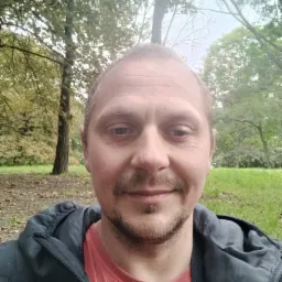 Mężczyzna z lekkim zarostem robiący selfie na tle parku z zieloną trawą i drzewami w jesiennej scenerii.