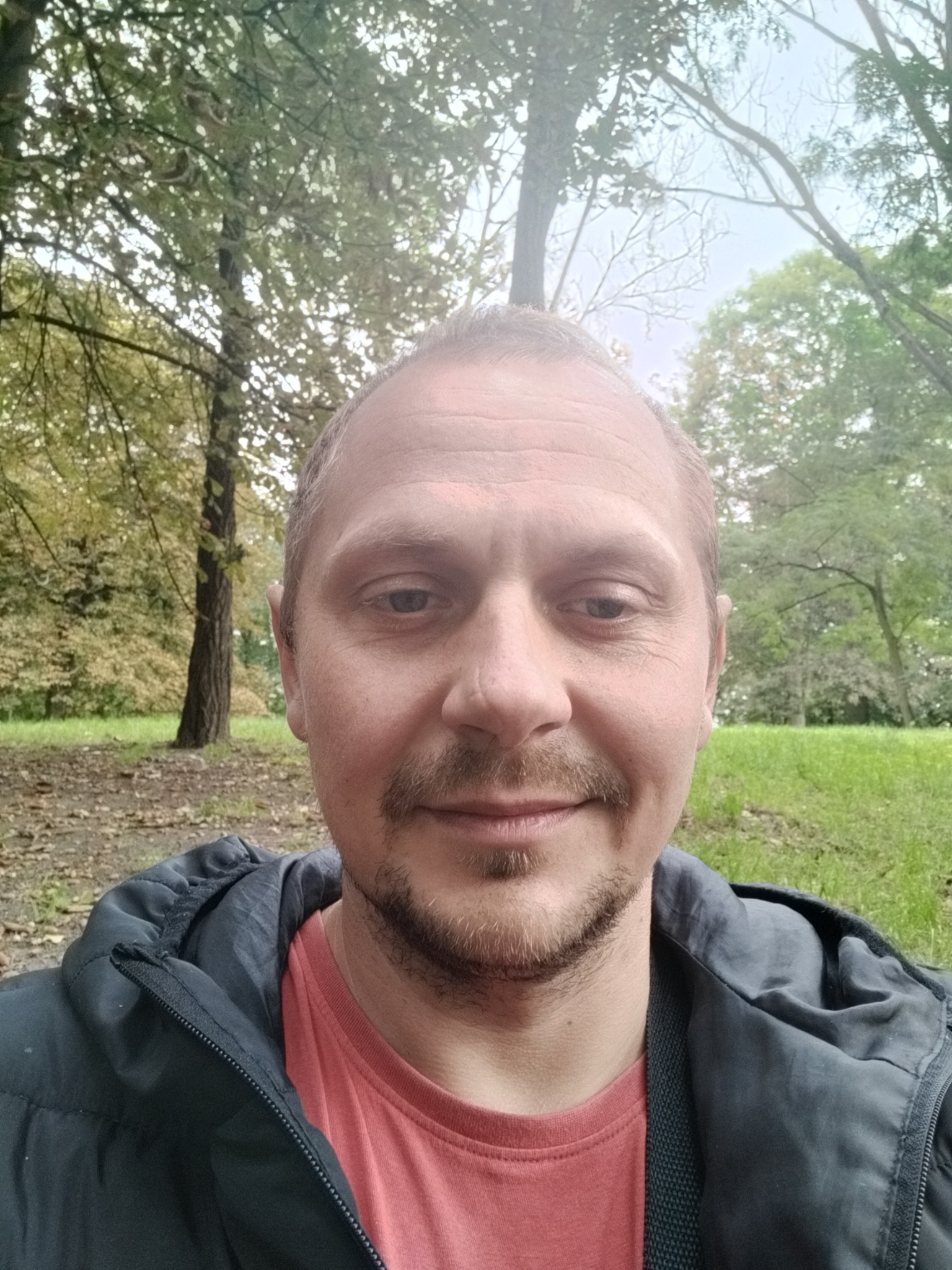 Mężczyzna z lekkim zarostem robiący selfie na tle parku z zieloną trawą i drzewami w jesiennej scenerii.