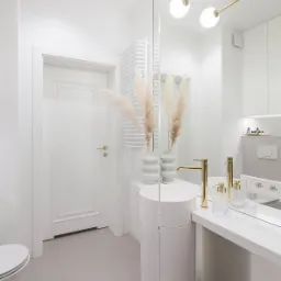 Jasna, minimalistyczna łazienka z okrągłą umywalką na postumencie, złotą armaturą, pampasami w dekoracyjnych wazonach i dużym lustrem.