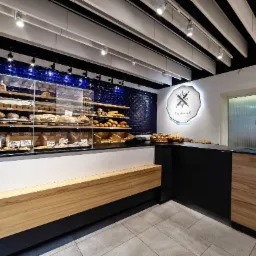 Wnętrze nowoczesnej piekarni z ladą z drewnianym frontem, ekspozycją pieczywa w szklanych gablotach na tle ściany z ciemnoniebieskich płytek oraz logo piekarni na ścianie.