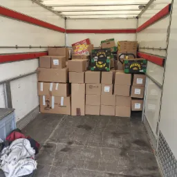 Wnętrze furgonetki załadowane kartonami, widoczne kartony z napisem 'Yellow Premium Bananas' i 'Kellogg's Corn Flakes', część ładunku zabezpieczona dodatkowymi opakowaniami.