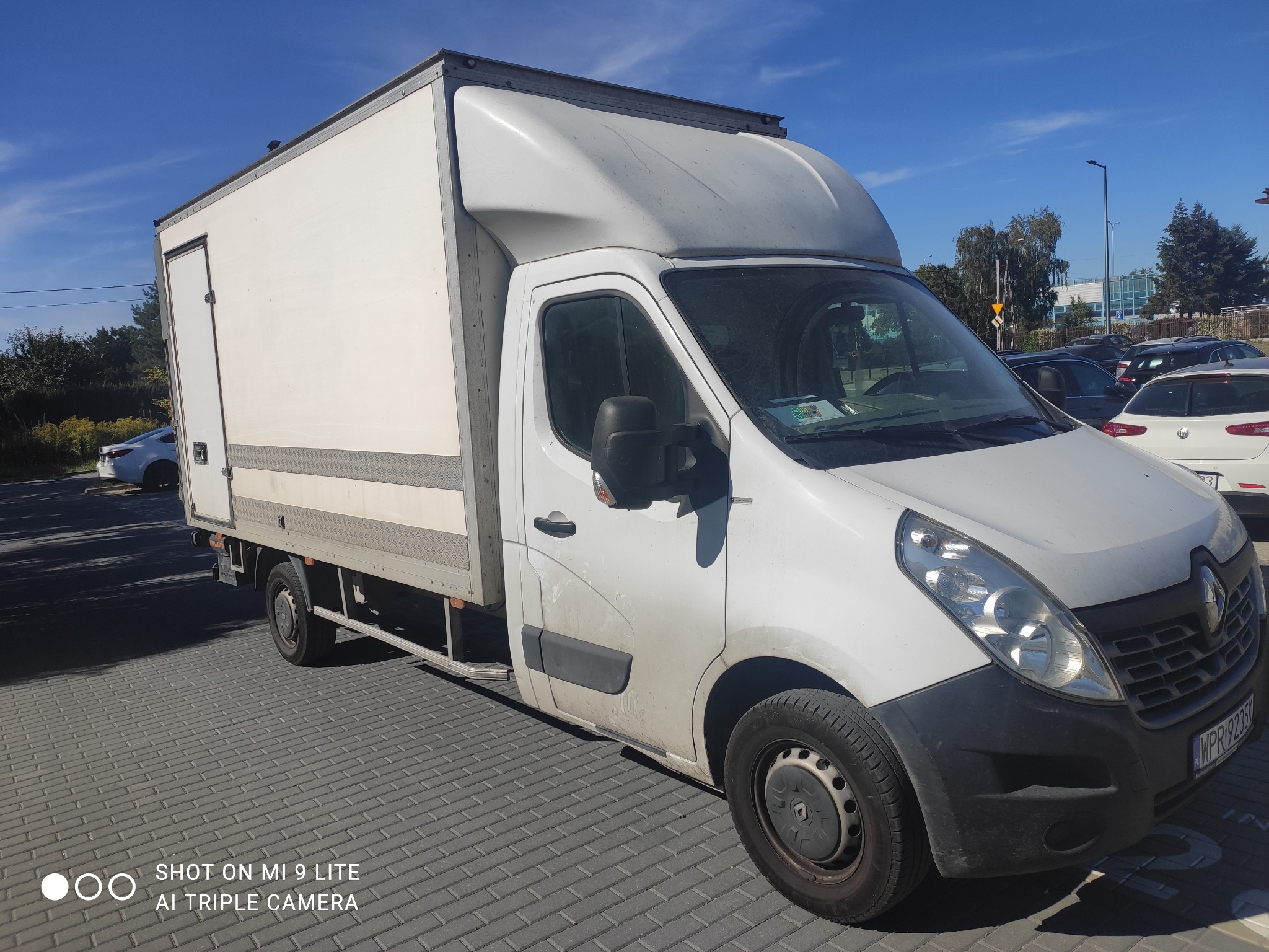 Biały samochód dostawczy Renault Master z zabudową kontenerową, zaparkowany na szarej kostce brukowej, widoczny numer rejestracyjny i otoczenie parkingu z innymi pojazdami w tle.