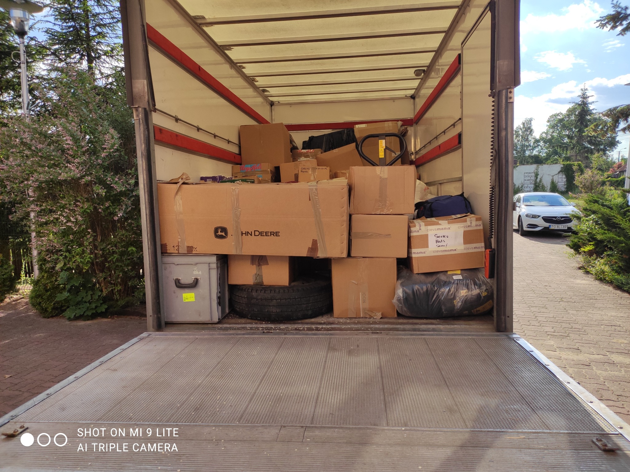 Wnętrze skrzyni ładunkowej wypełnione kartonami, w tym karton z logo John Deere, oraz opona samochodowa, widoczne elementy wyposażenia i pakunki, na zewnątrz samochód osobowy i zieleń.