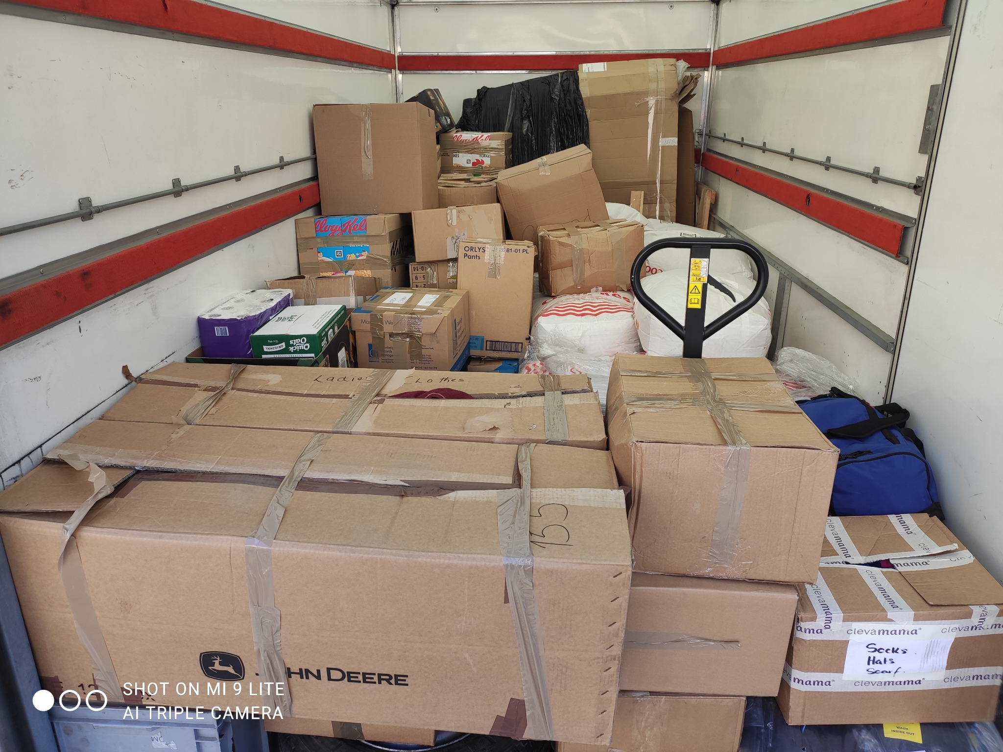 Wnętrze furgonetki wypełnione kartonami różnej wielkości i zawartości, niektóre z oznaczeniami 'Ladies Clothes' i 'HN Deere', z widocznym wózkiem paletowym w tle.