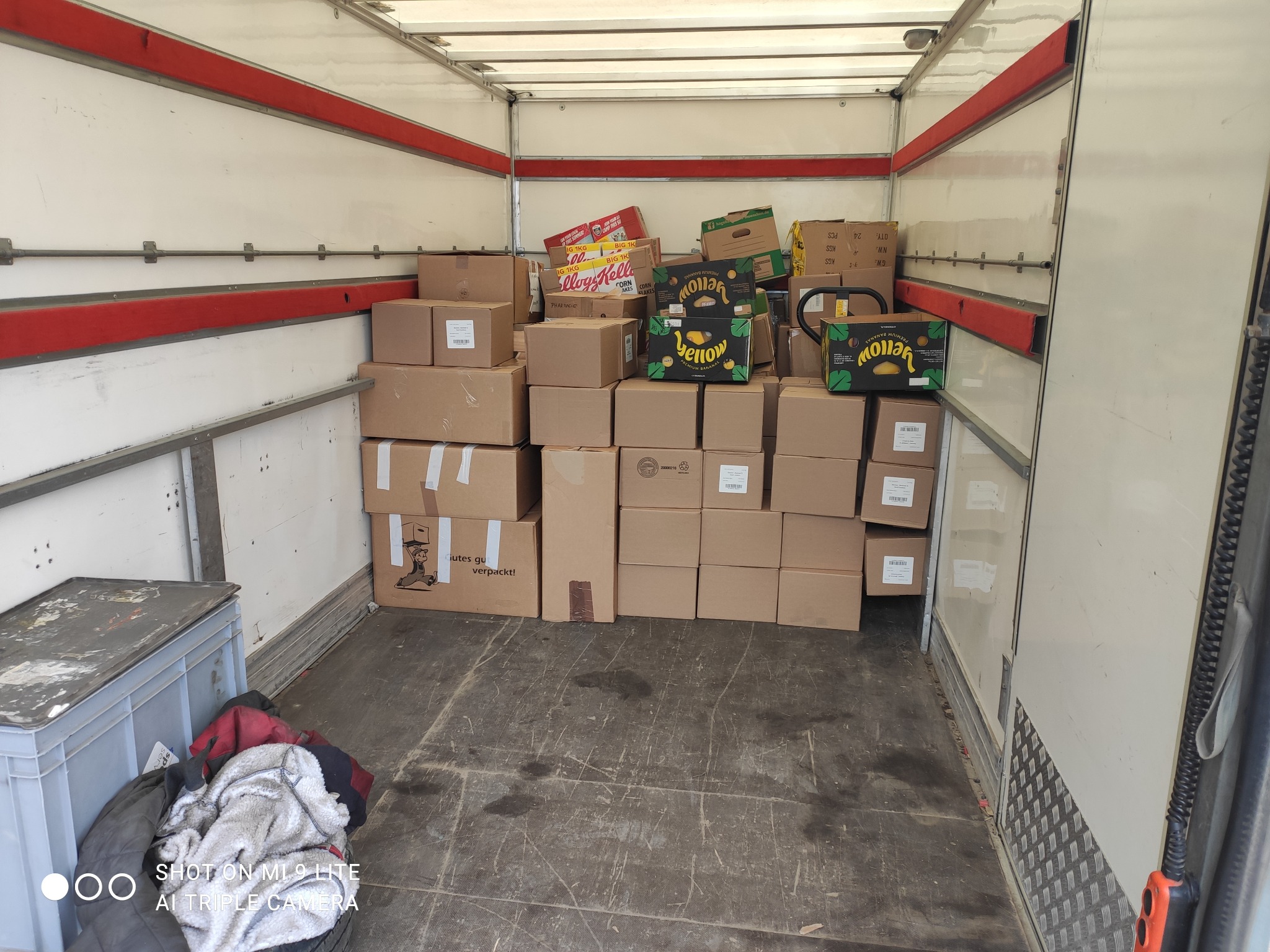 Wnętrze furgonetki załadowane kartonami, widoczne kartony z napisem 'Yellow Premium Bananas' i 'Kellogg's Corn Flakes', część ładunku zabezpieczona dodatkowymi opakowaniami.