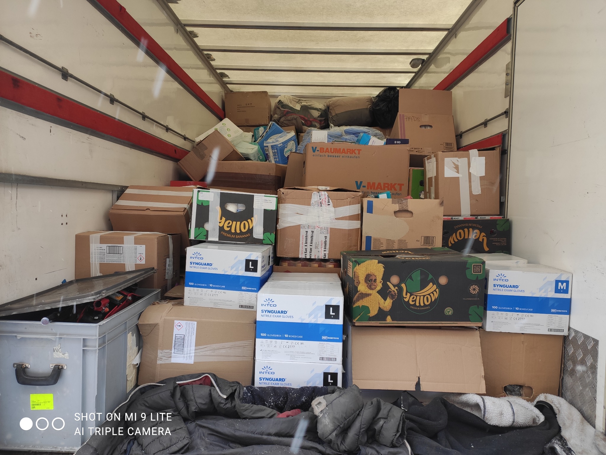 Wnętrze furgonetki wypełnione kartonami różnej wielkości i zawartości, widoczne pudełka z rękawicami ochronnymi i bananami, zabezpieczone do przewozu.