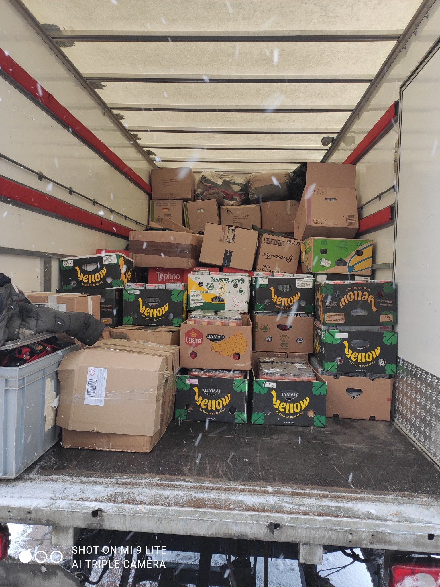 Wnętrze częściowo załadowanej ciężarówki z kartonami, niektóre z oznaczeniem 'Yellow Premium Bananas', widoczne padające płatki śniegu.