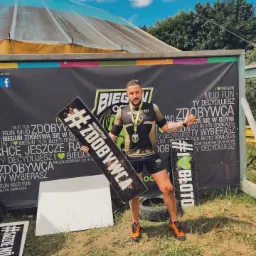 Uśmiechnięty mężczyzna z medalem, ubrudzony błotem po biegu OCR, trzymający tabliczki z hasztagami #ZDOBYWCA i #BLOTO na tle baneru reklamowego biegu.