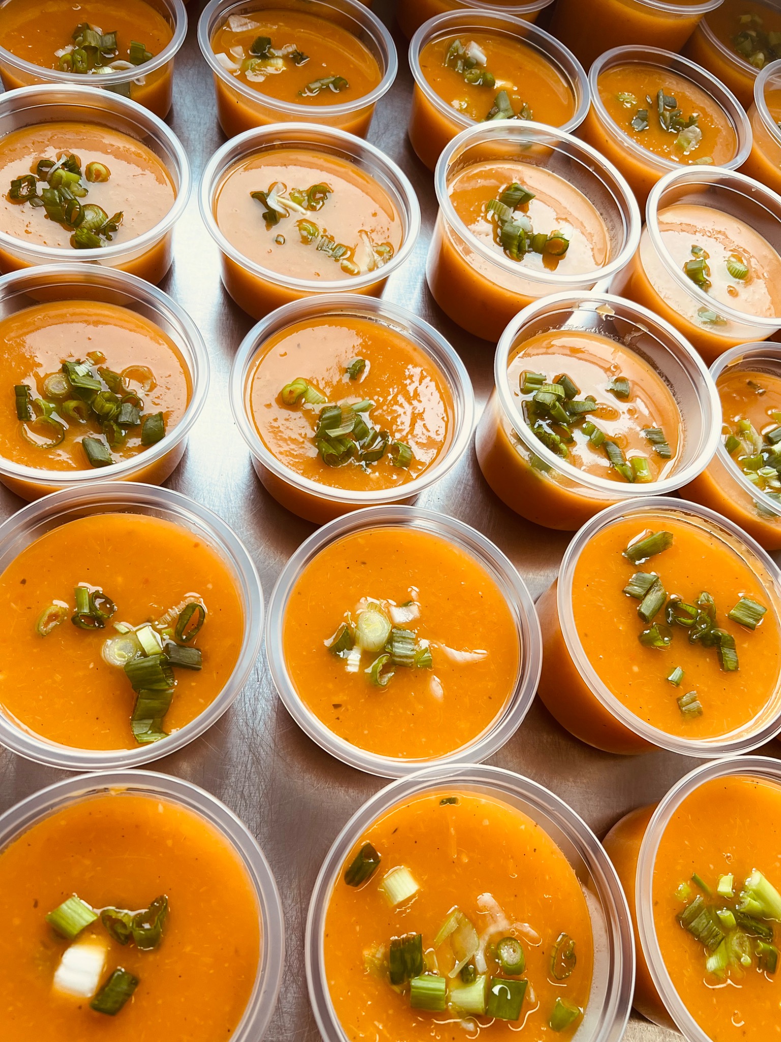 Porcje kremowej pomarańczowej zupy w plastikowych pojemnikach, posypane świeżym szczypiorkiem, ułożone rzędami na metalowej powierzchni.