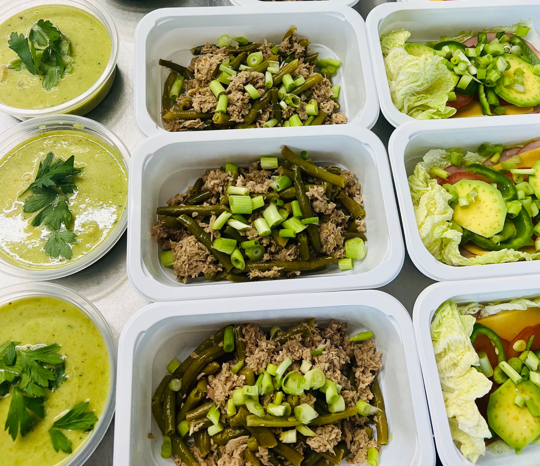 Zestaw dietetycznych posiłków w plastikowych pojemnikach: kremowa zupa z natką pietruszki, tuńczyk z fasolką szparagową i zielonym szczypiorkiem, sałatka z awokado, sałaty, papryki i szynki.