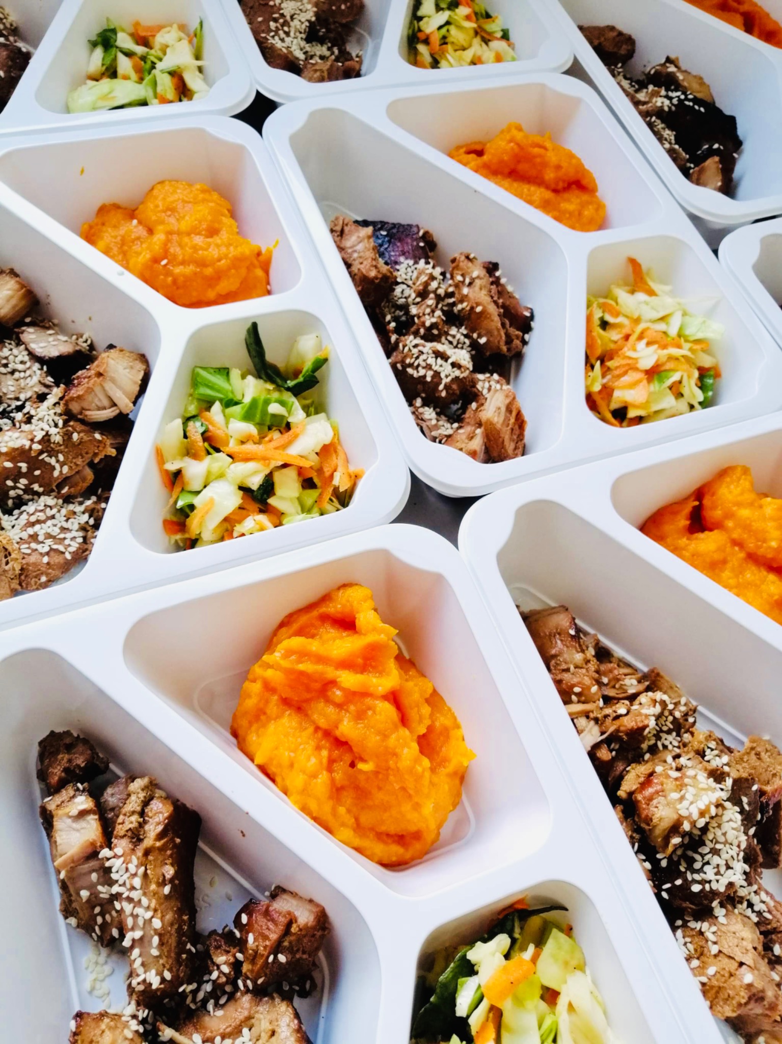 Zestaw dań w białych, podzielonych pojemnikach: pieczone mięso posypane sezamem, puree z batatów i surówka z kapusty, marchewki i ogórka.