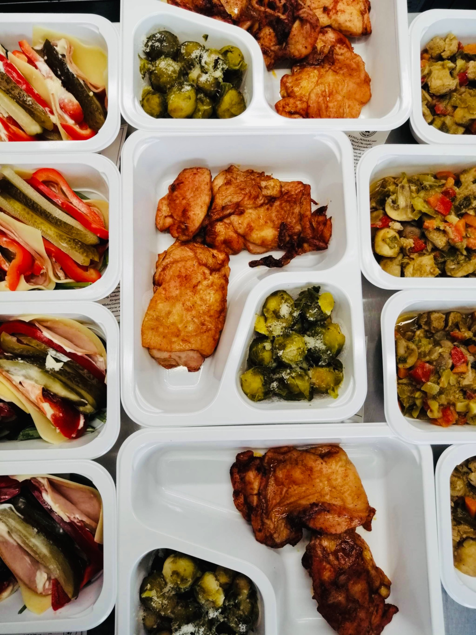 Zestaw gotowych posiłków w białych, plastikowych pojemnikach: pieczony kurczak, brukselka z parmezanem, surówka z warzyw, sałatka z ogórków, papryki i szynki.