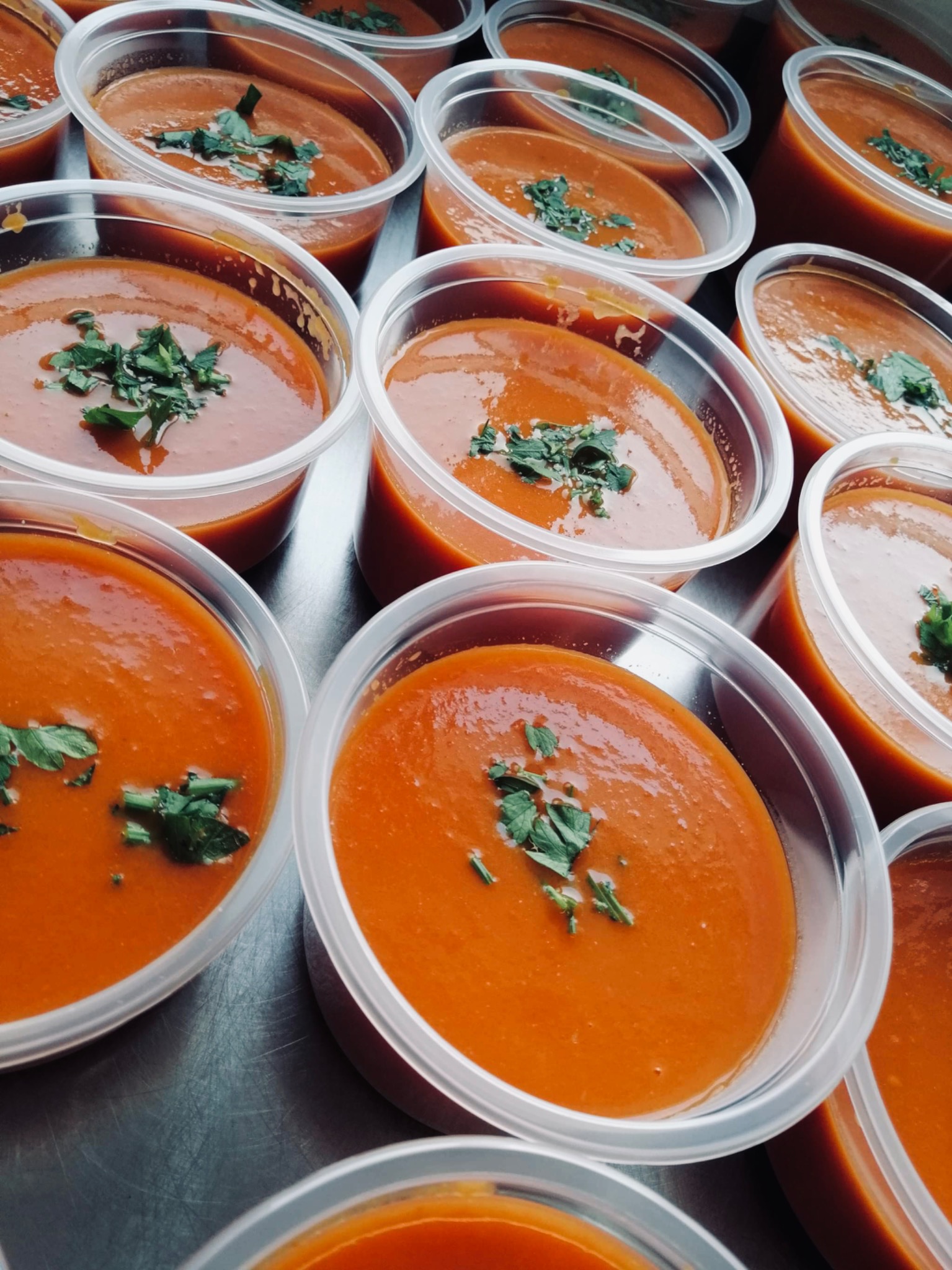 Porcje kremu pomidorowego w przezroczystych pojemnikach z dodatkiem świeżej zielonej pietruszki, ułożone rzędami na metalowej powierzchni.