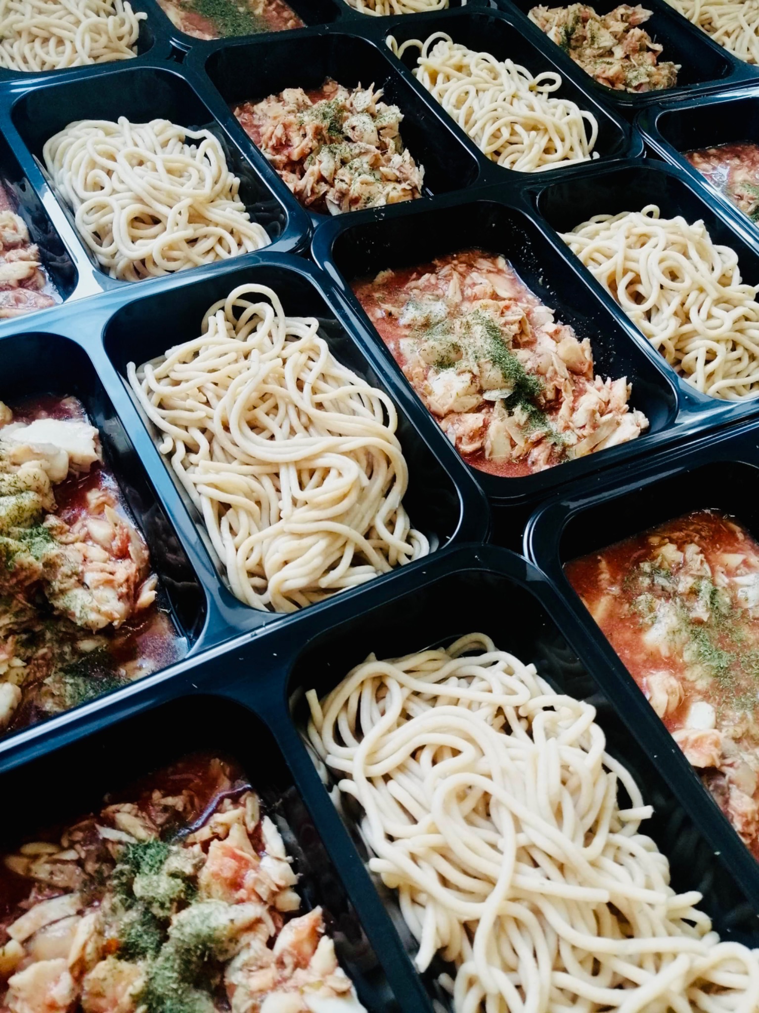 Porcje spaghetti z sosem pomidorowym i ziołami w czarnych, plastikowych pojemnikach, ułożone w regularny wzór, widoczne z góry.