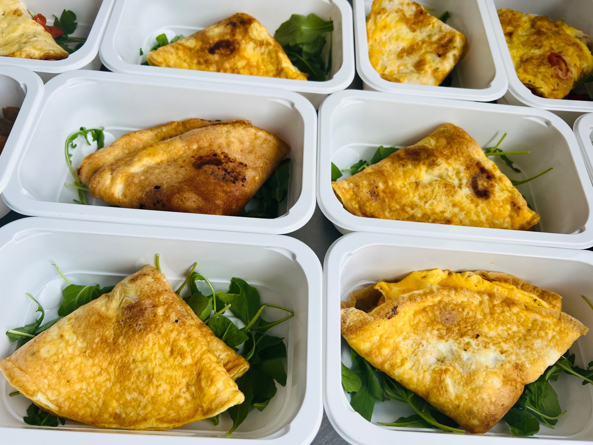 Owsiane omlety z rukolą w białych, plastikowych pojemnikach, widoczne z góry, gotowe do transportu.