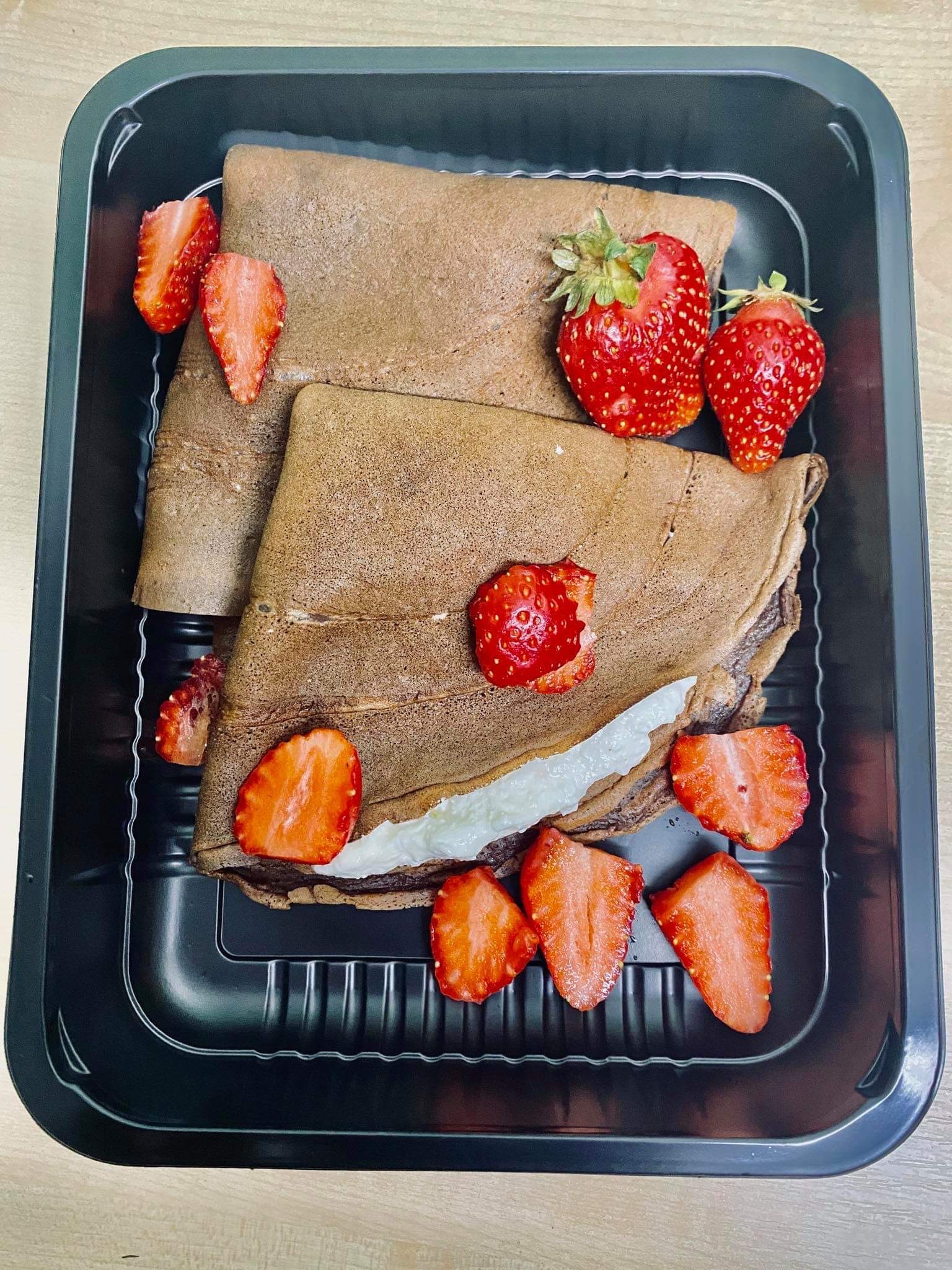 Dwa czekoladowe naleśniki z kremem w czarnym pudełku, udekorowane połówkami świeżych truskawek.