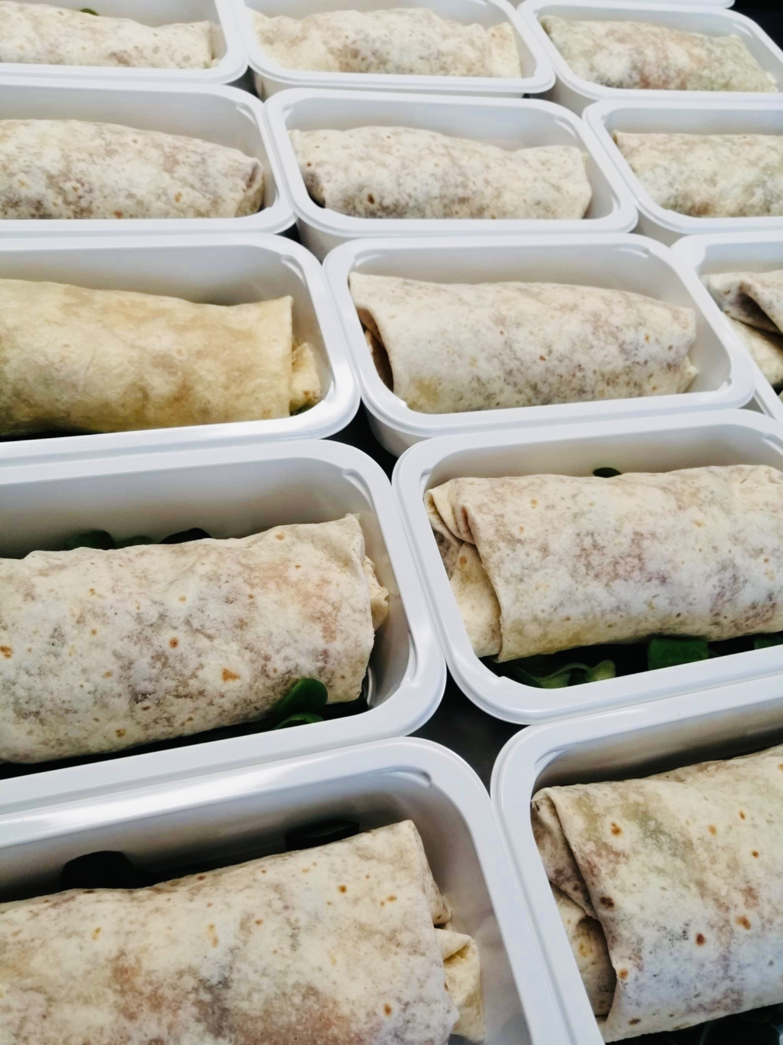 Rząd plastikowych pojemników z zawiniętymi tortillami z nadzieniem i zielonymi liśćmi, prawdopodobnie przygotowanych do transportu.