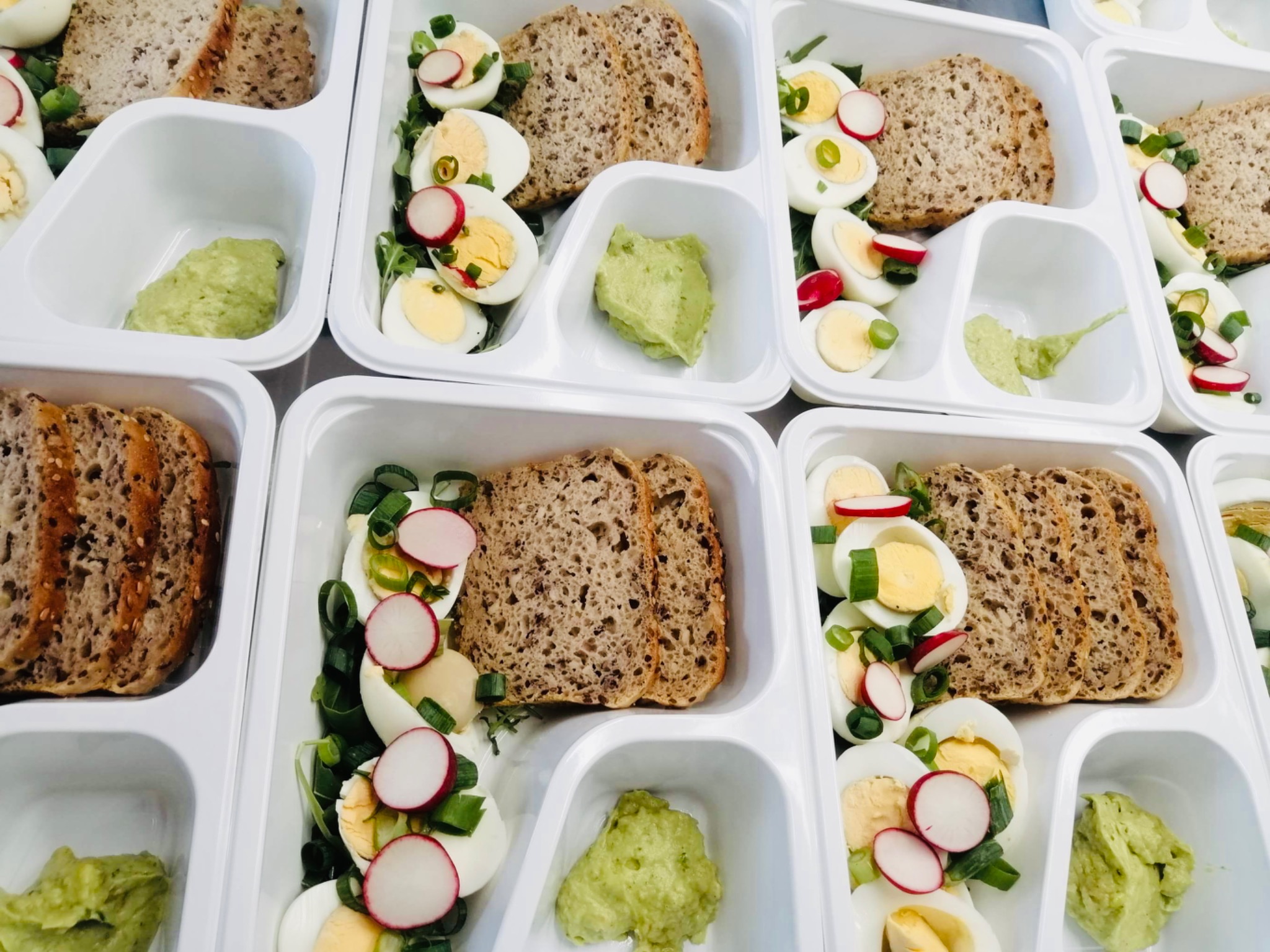 Porcje dietetycznego posiłku w białych, plastikowych pojemnikach: kromki ciemnego pieczywa, jajka na twardo z rzodkiewką i szczypiorkiem, oraz porcja zielonego guacamole.
