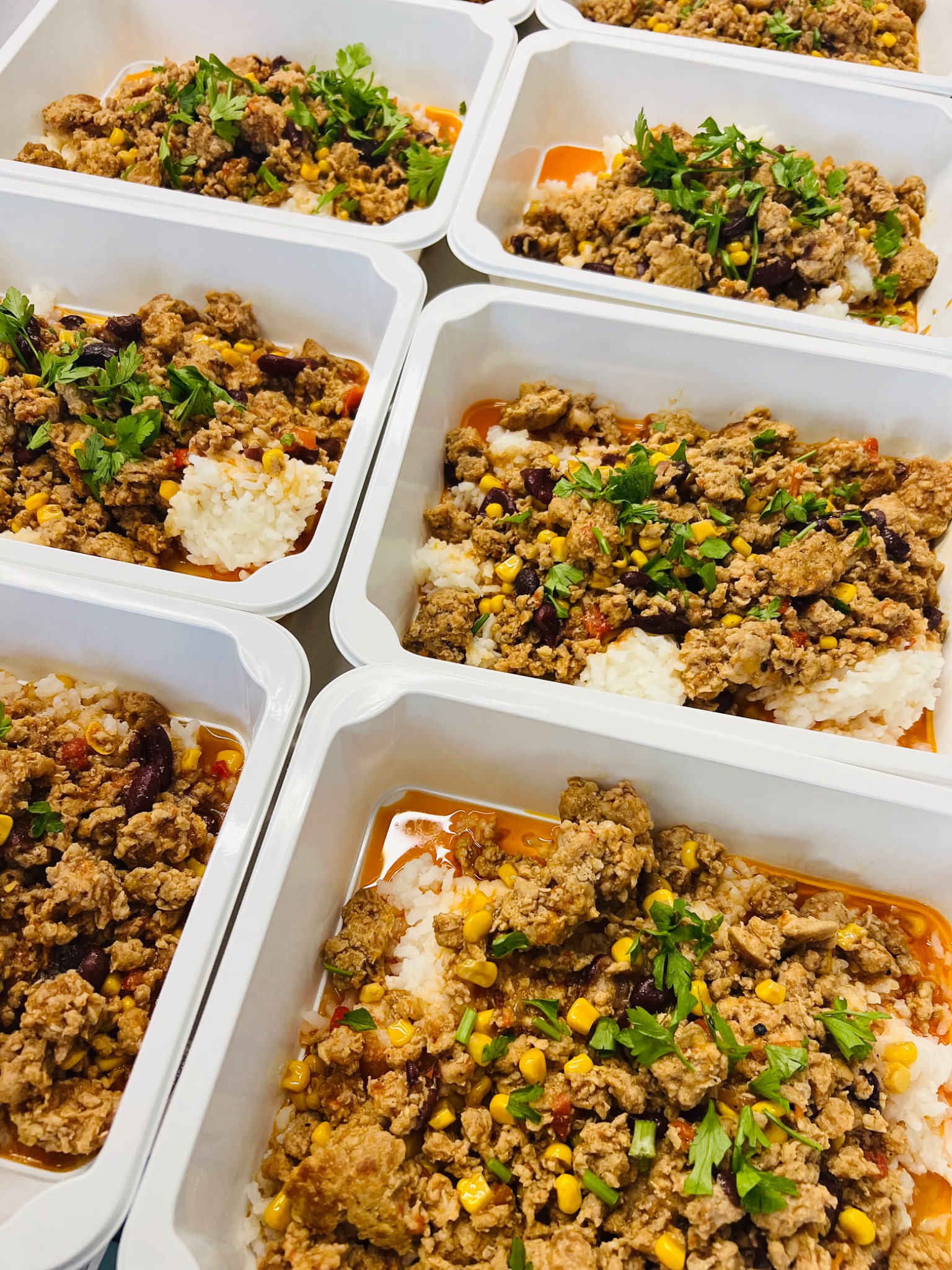 Gotowe porcje chili con carne z ryżem w białych pojemnikach, posypane świeżą kolendrą i kukurydzą, widok z góry.