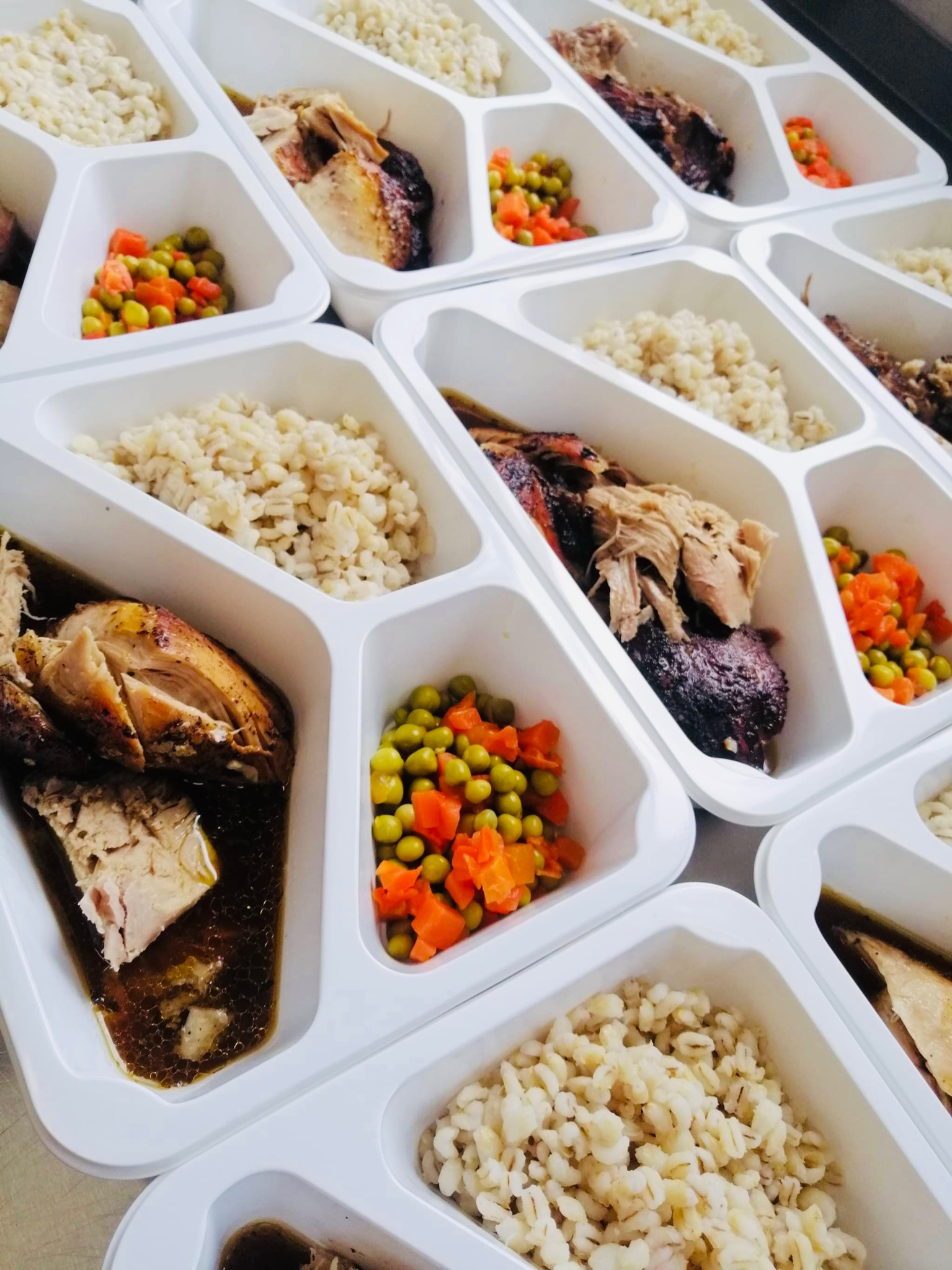 Porcje cateringu dietetycznego w białych, plastikowych pojemnikach z trzema przegródkami: pieczone mięso, kasza jęczmienna i gotowana marchewka z groszkiem.