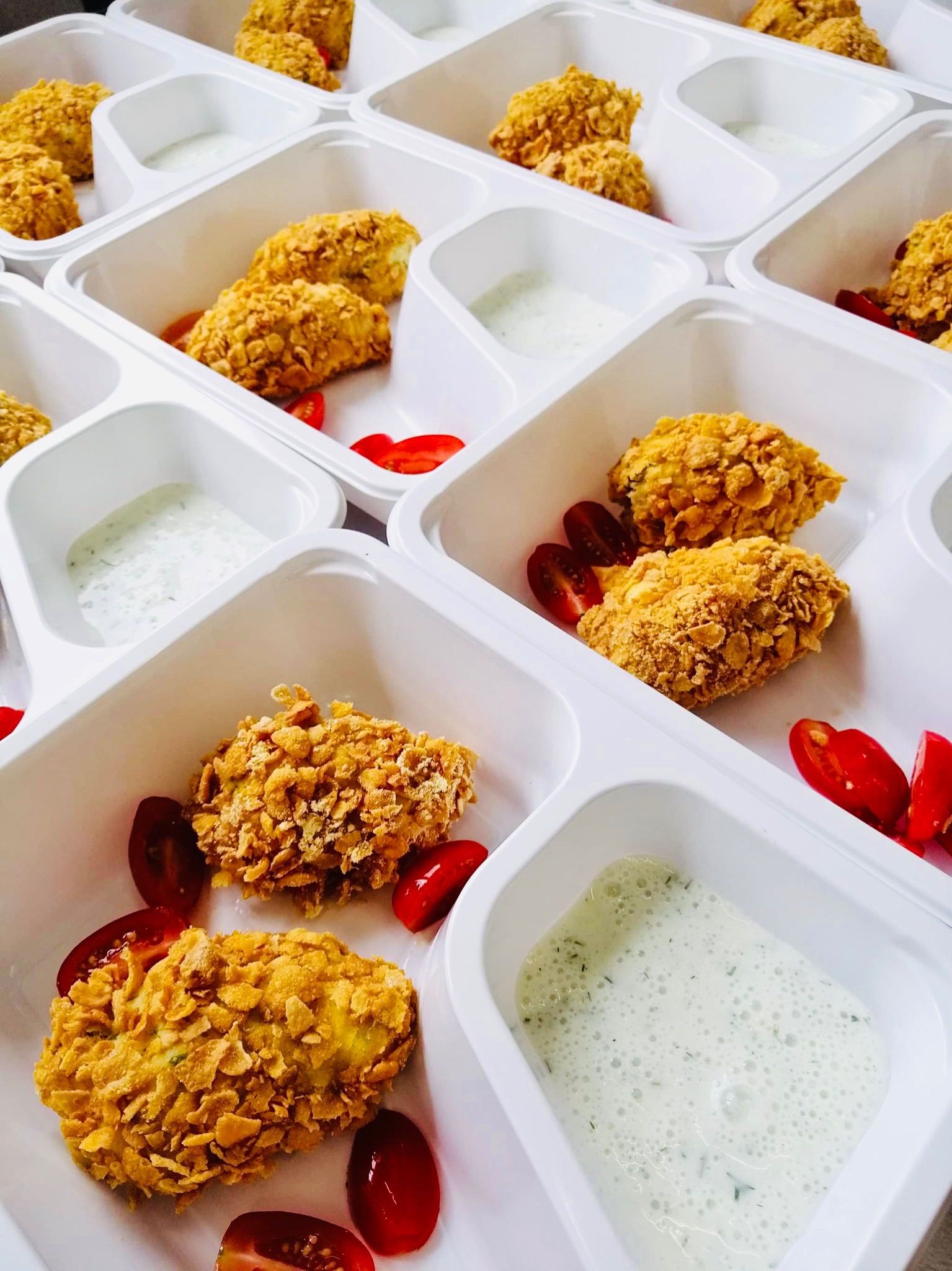 Porcje nuggetsów w panierce z płatków kukurydzianych, podane z sosem ziołowym i pomidorkami koktajlowymi w białych, plastikowych pojemnikach cateringowych od firmy z Tucholi.