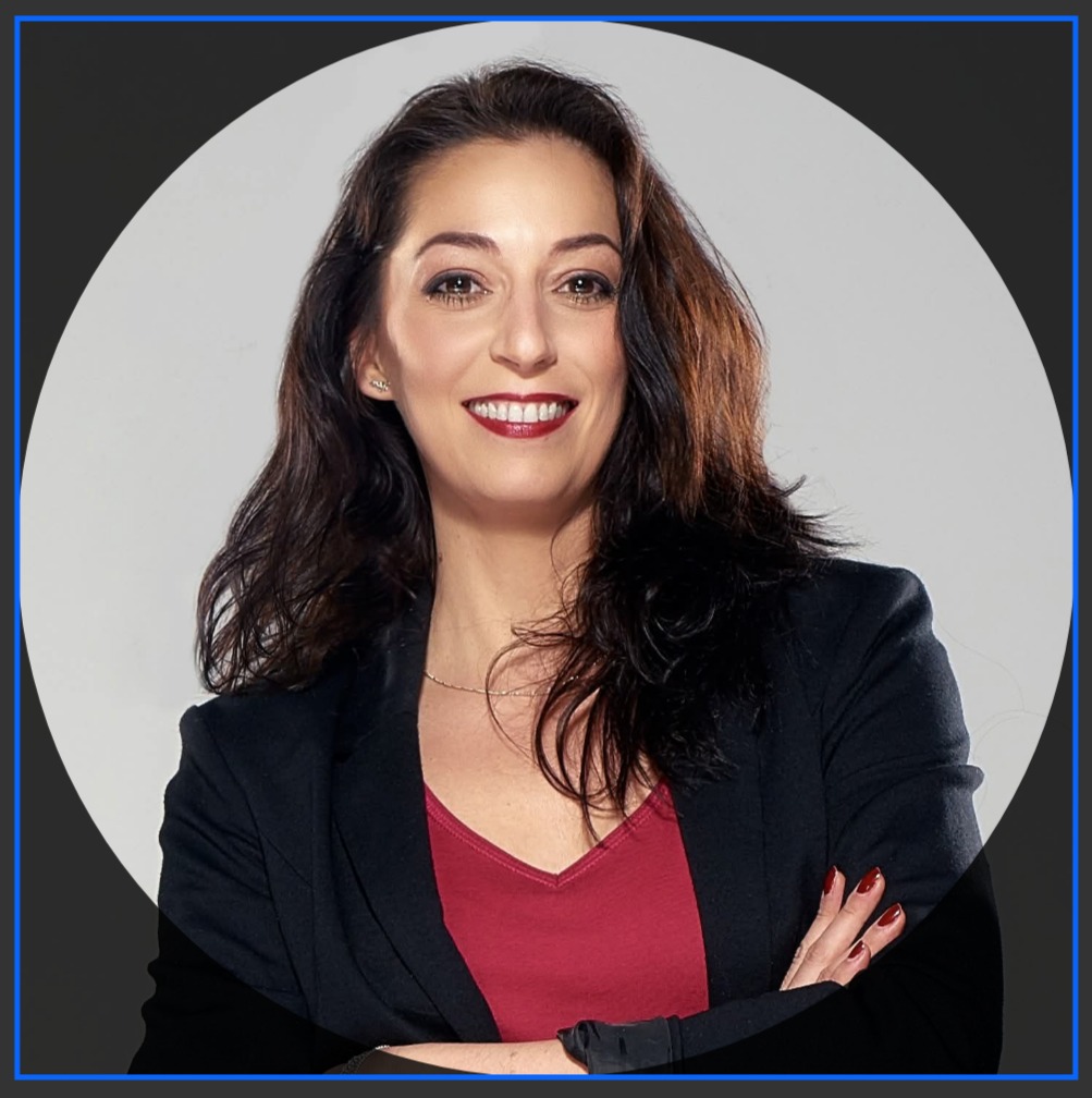 Portret uśmiechniętej kobiety w czarnej marynarce i czerwonej bluzce, z rękami skrzyżowanymi na piersiach, na jasnym tle w okręgu.