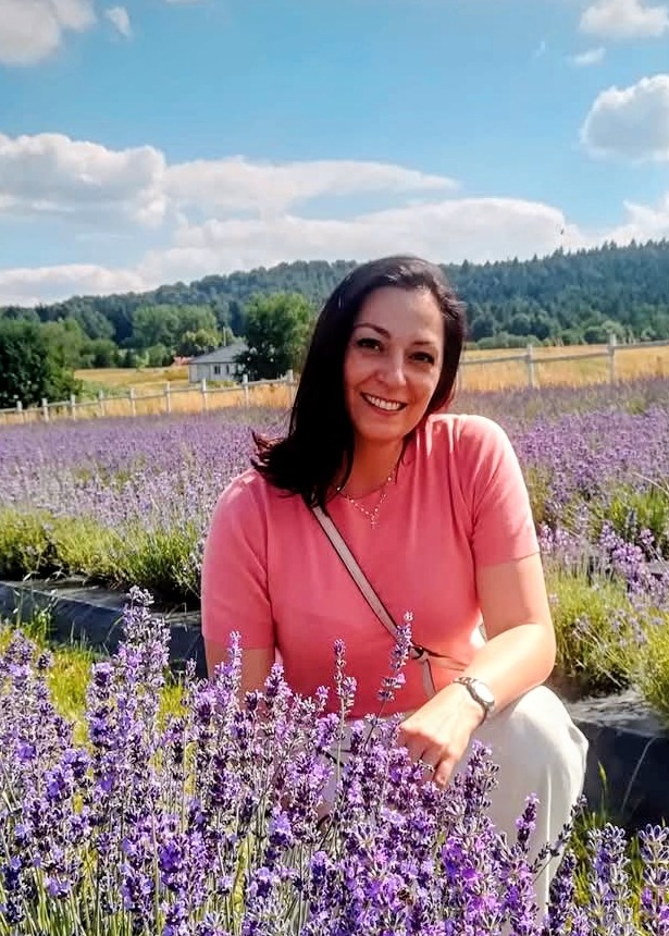 Uśmiechnięta kobieta w różowej koszulce siedzi na polu lawendy w słoneczny dzień. W tle zielone wzgórza i błękitne niebo z chmurami.