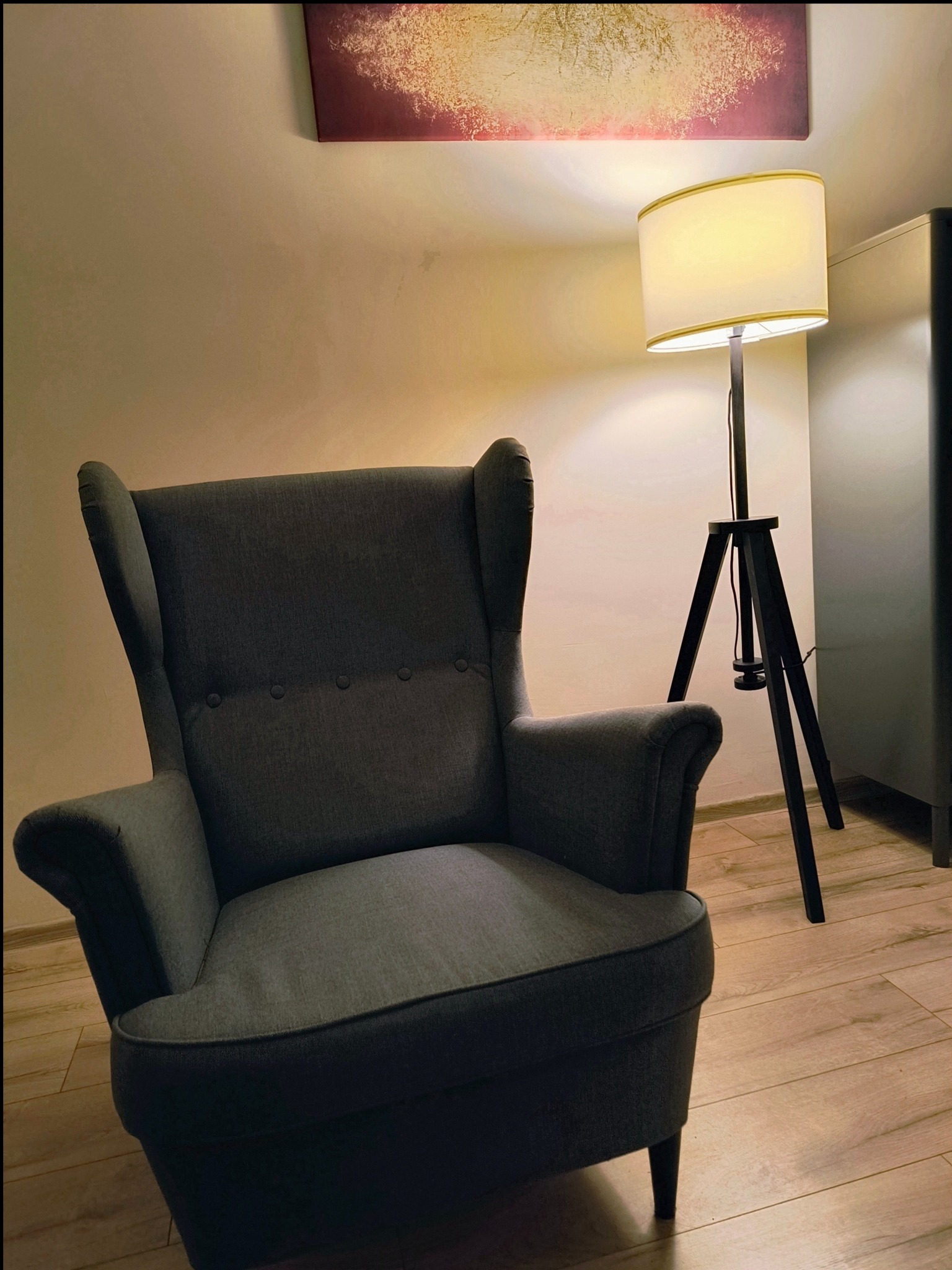 Przytulny, zaciszny kącik z szarym fotelem typu uszak, lampą stojącą na trójnogu i abstrakcyjnym obrazem na ścianie, tworzący relaksującą atmosferę.