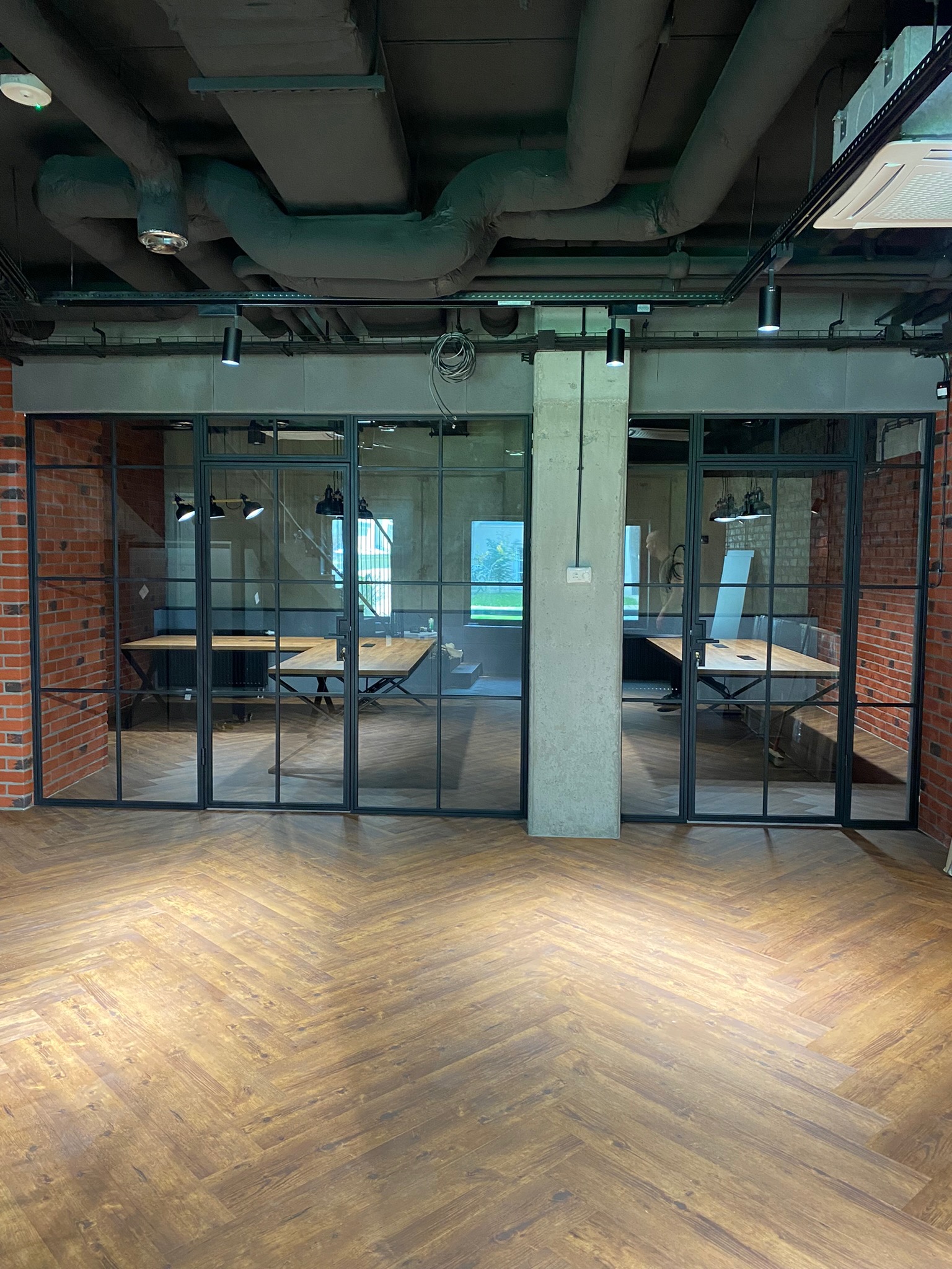 Przeszklona ściana działowa z czarnymi ramami oddzielająca przestrzeń biurową, widoczna ceglana ściana po lewej stronie i drewniana podłoga ułożona w jodełkę.
