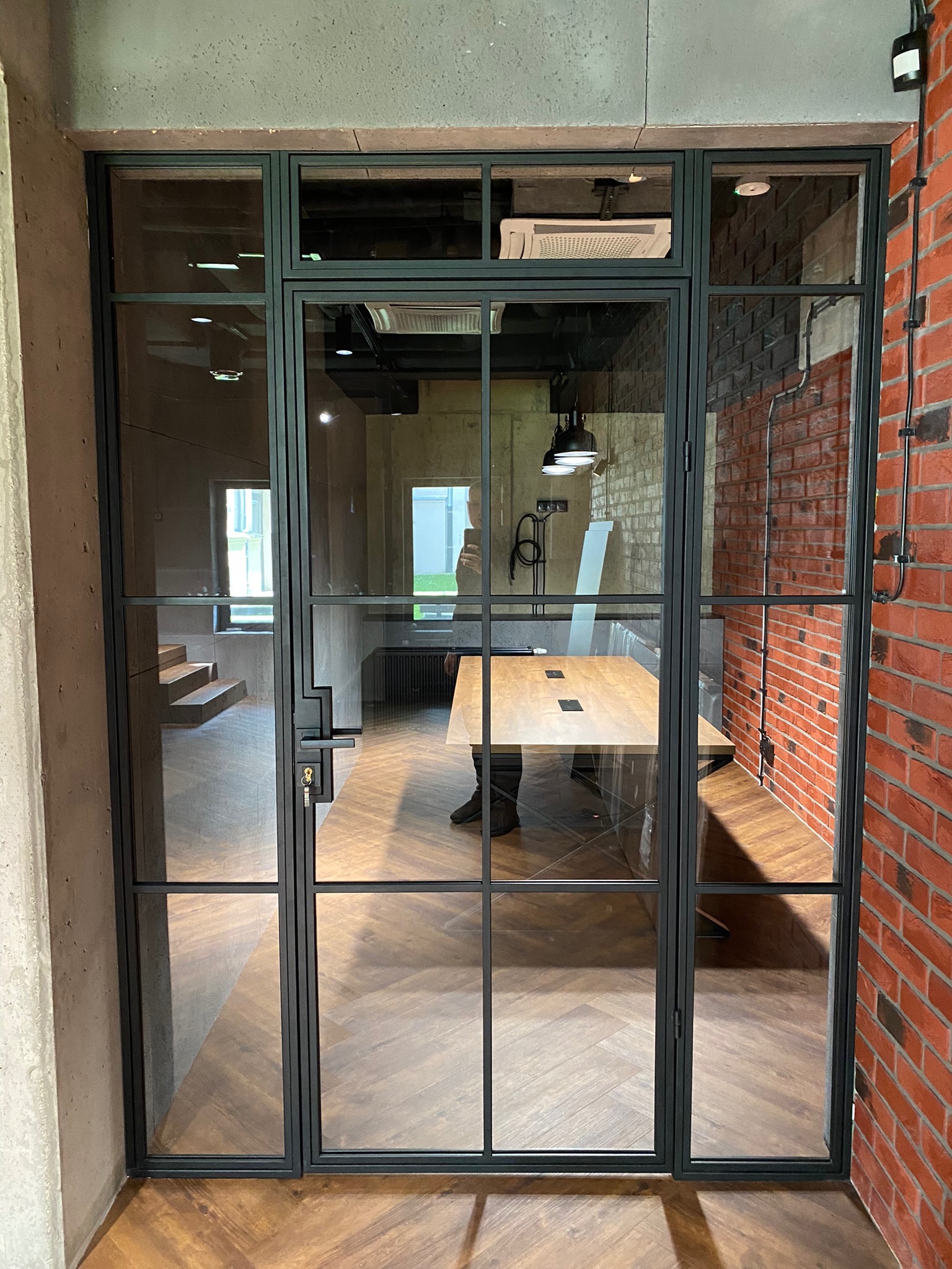 Szklane drzwi wewnętrzne z czarnymi szprosami oddzielające biuro z ceglaną ścianą i drewnianą podłogą w jodełkę od klatki schodowej.