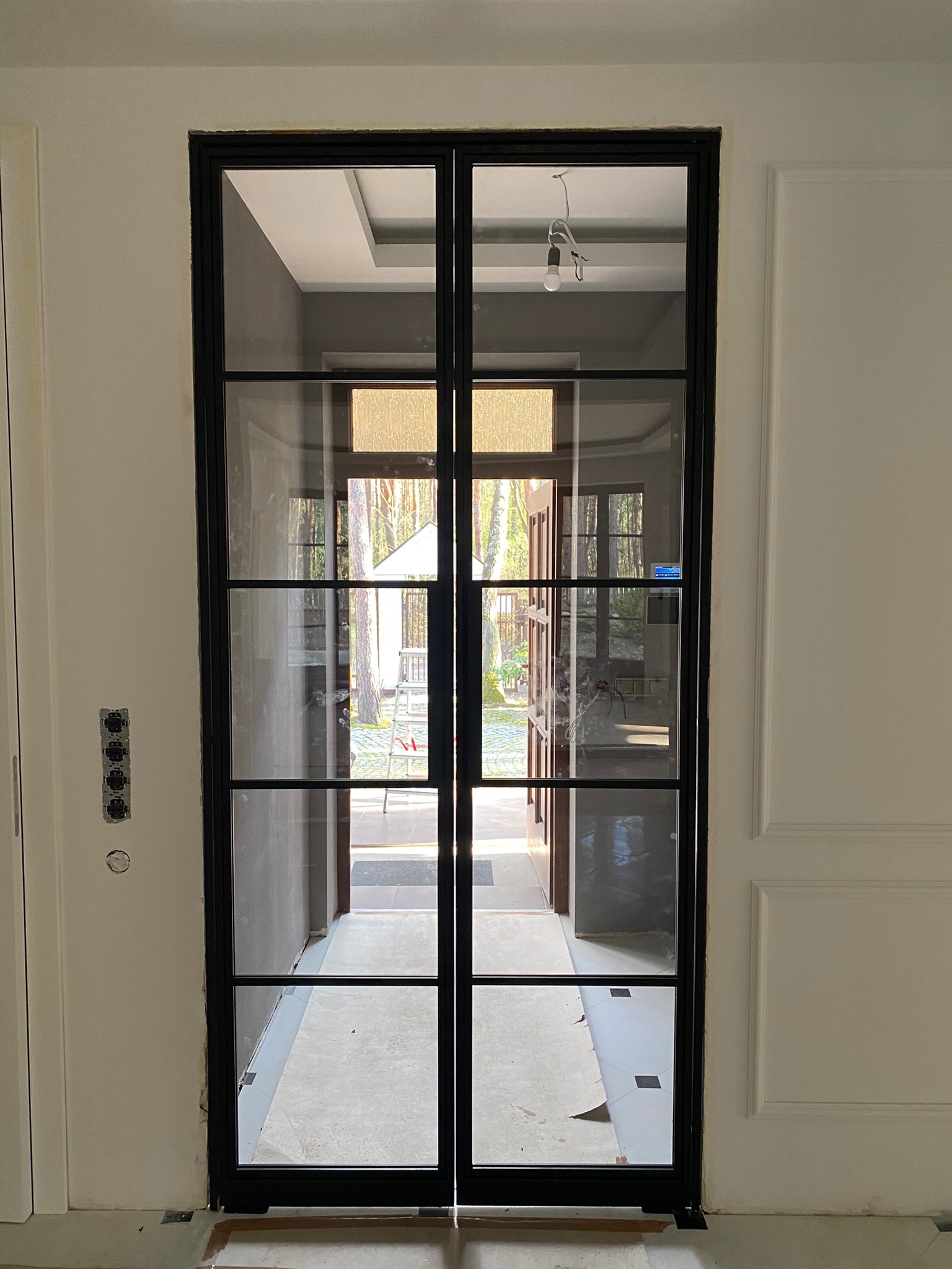 Dwuskrzydłowe, czarne drzwi z szybami w stylu industrialnym, widok na wnętrze domu i ogród, surowy stan wykończenia podłogi.