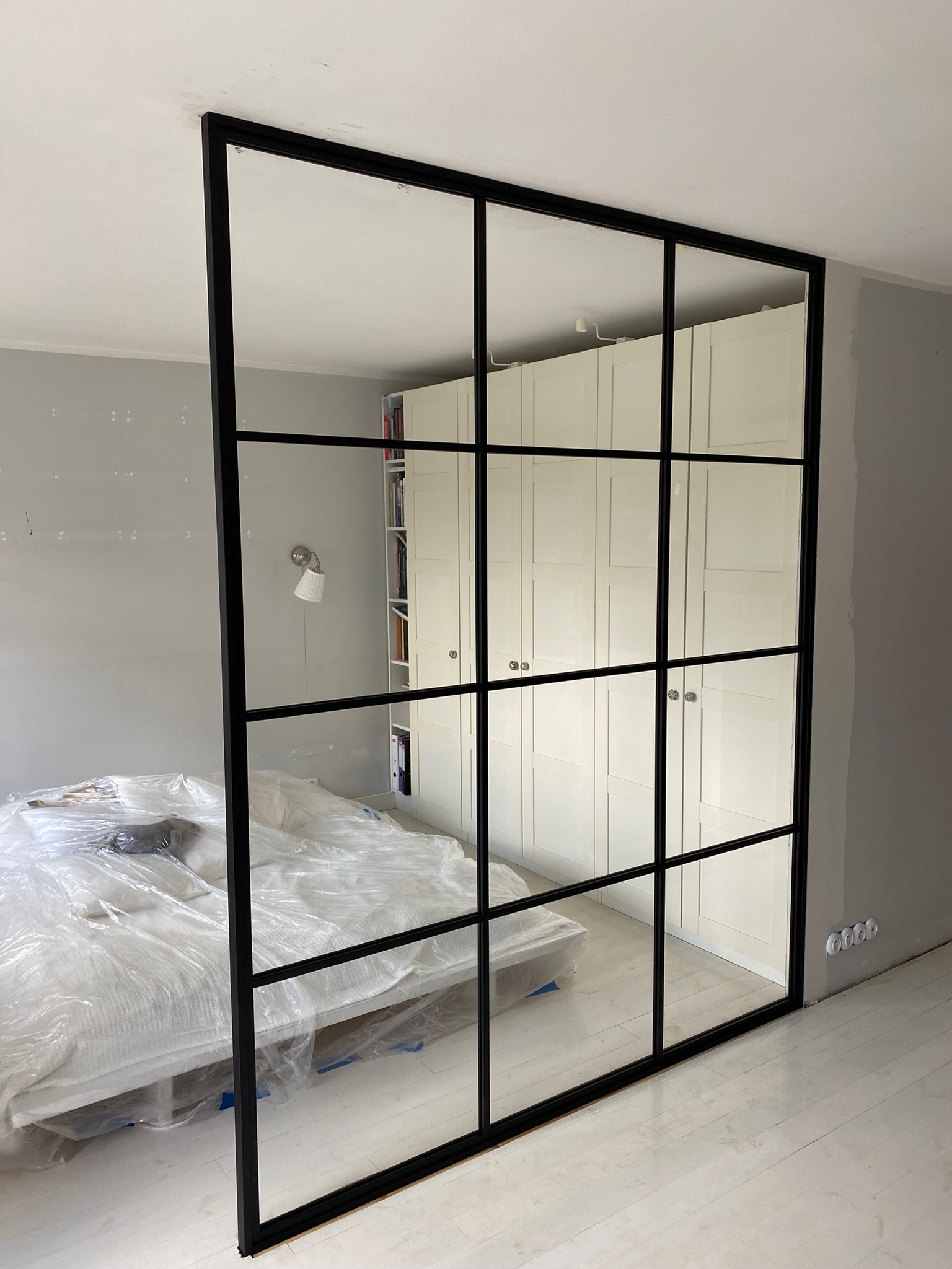 Czarna, stalowa ścianka działowa ze szklanymi panelami oddzielająca sypialnię z łóżkiem przykrytym folią ochronną od części z białą zabudową meblową.